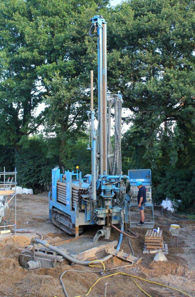 Blauwe boorinstallatie op rupsbanden maakt een verticale boring in een tuin, met boorbuizen op het chassis