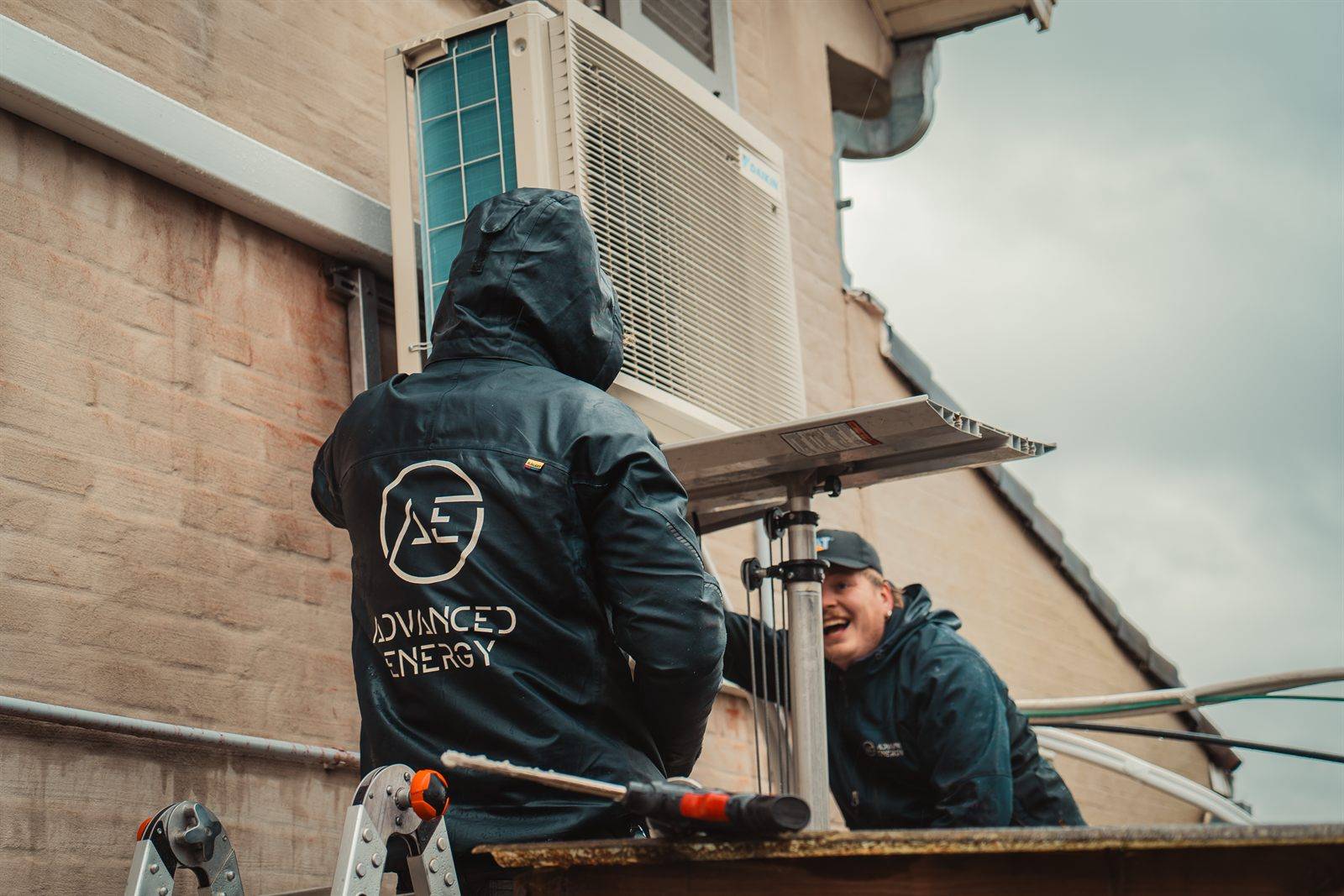 Twee techniekers van Advanced Energy plaatsen de buitenunit van een warmtepomp tegen een gevel