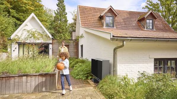 Daikin Altherma 4 lucht-water warmtepomp buitenunit tegen de gevel van een bestaande woning met pannendak, met een kind en een volwassene in de tuin